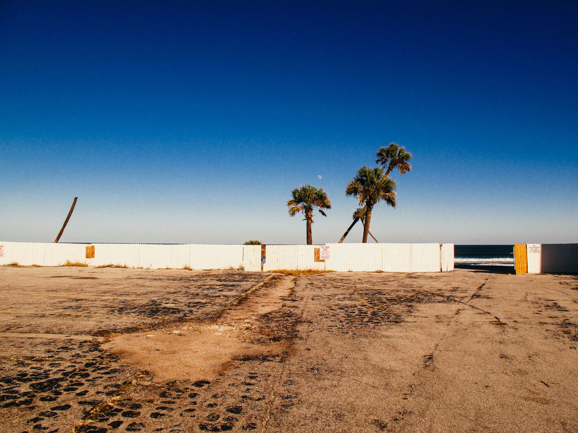 Empty cracked concrete parking lot facing a white wooden fence with openings showing palm trees and ocean waves beyond under a clear blue sky.