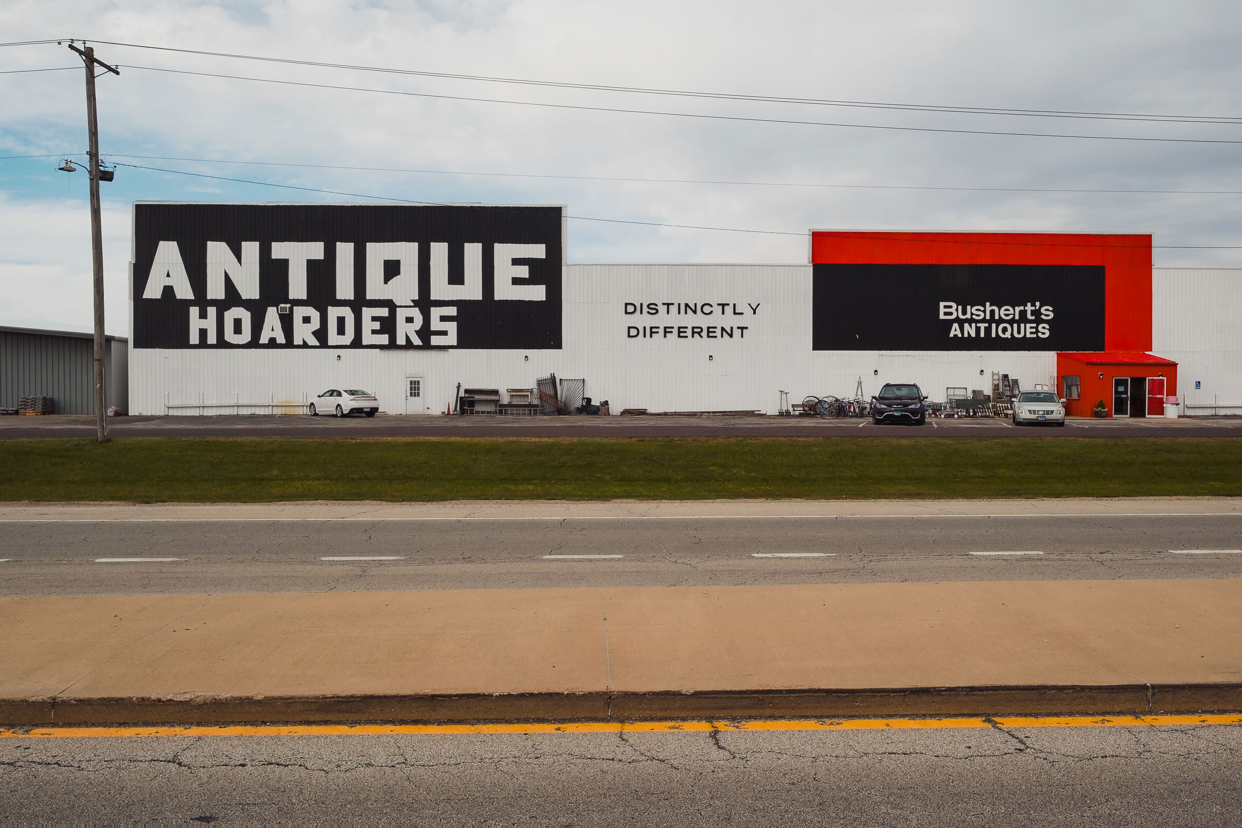 Wide exterior view of a large antique store building with three distinct sections: a black sign with large white letters reading 'ANTIQUE HOARDERS,' white wall with black letters saying 'DISTINCTLY DIFFERENT,' and a red-accented section with a black sign that says 'Bushert's ANTIQUES.' Several cars are parked in front, and various antiques are displayed outside the building.