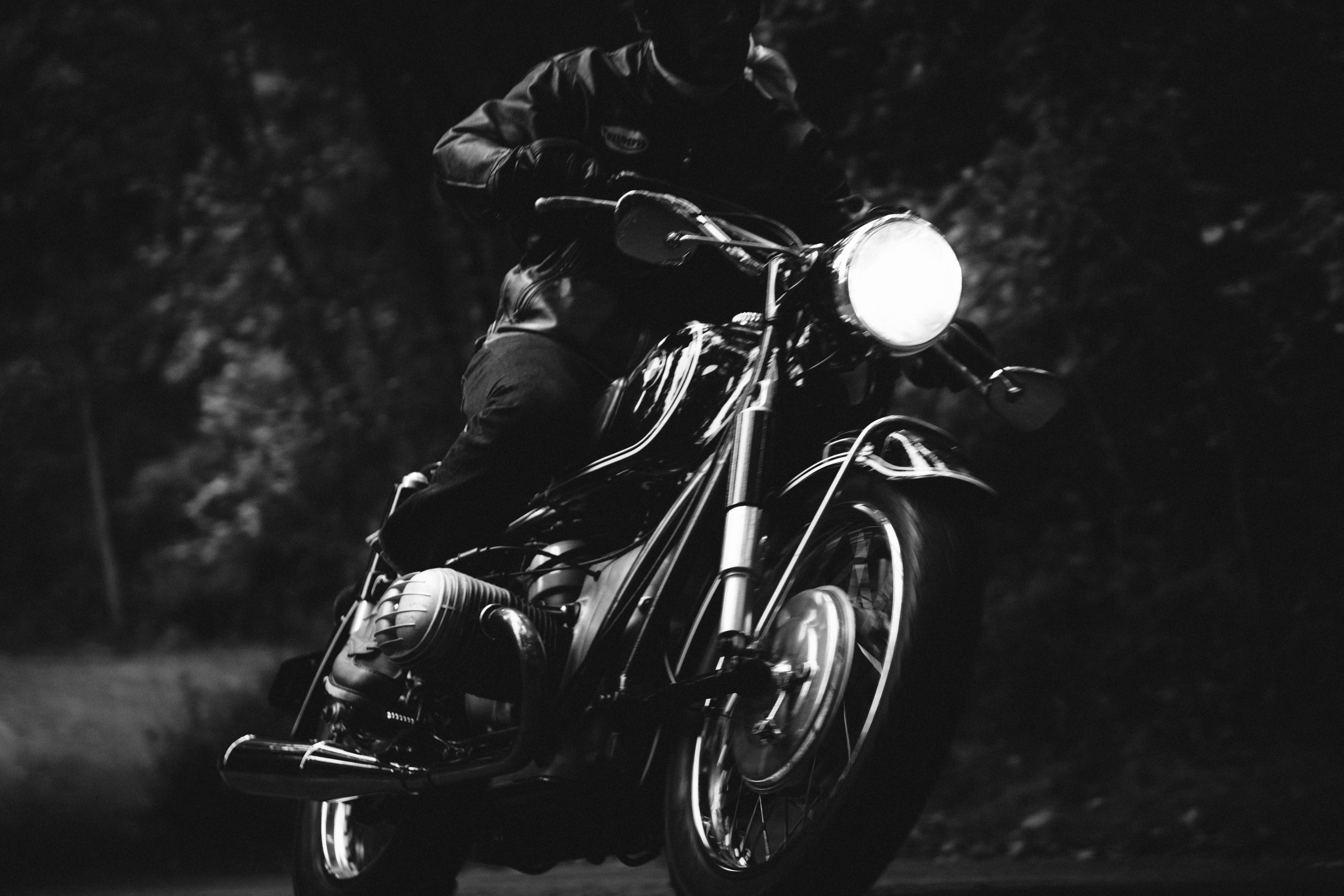 Black and white image of a person wearing a leather jacket and gloves riding a classic motorcycle with its front headlight on, surrounded by blurred trees in the background.
