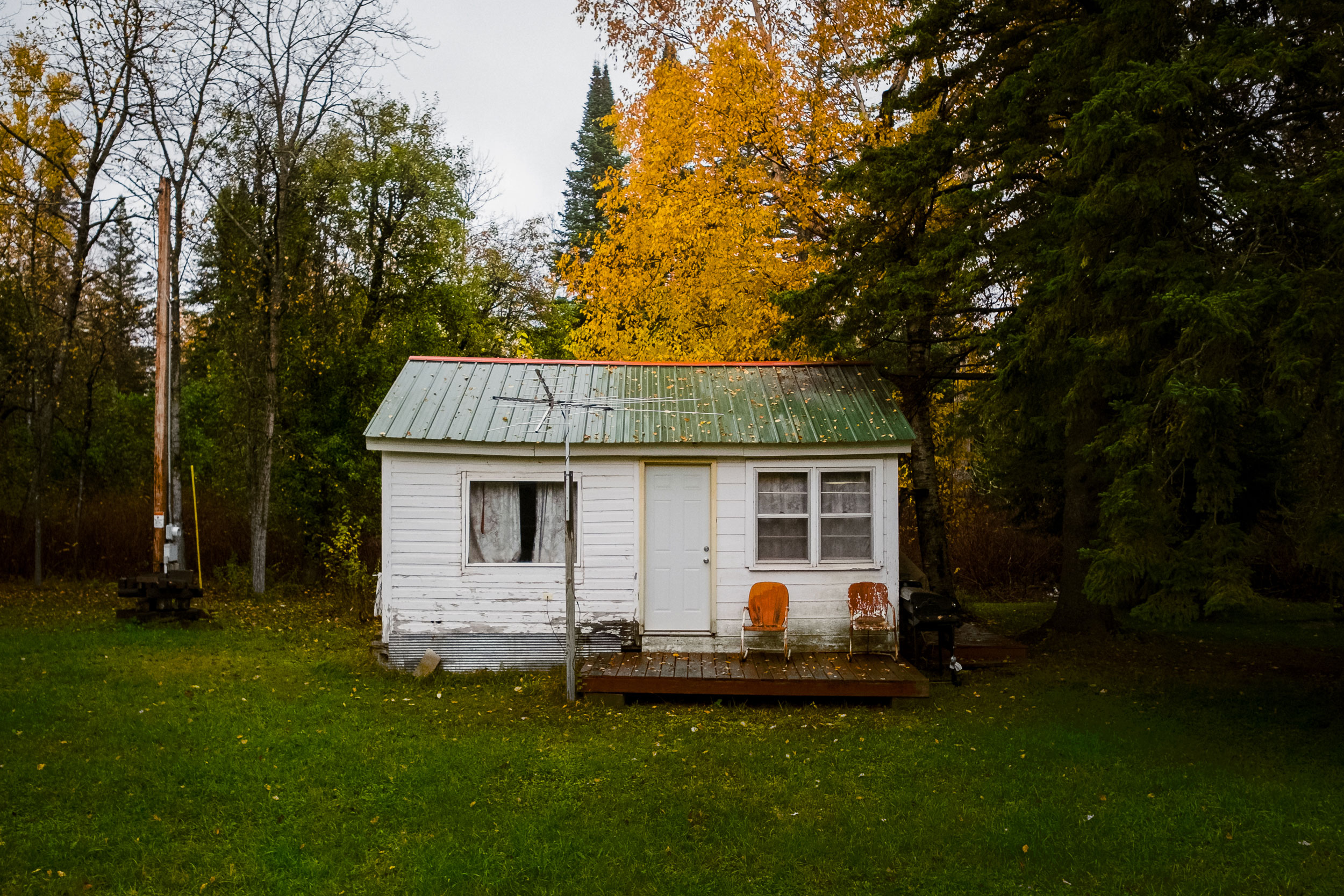 Small white wooden cabin with a green metal roof and a single door centered between two windows, surrounded by green grass and autumn trees with yellow leaves, with two orange chairs on the front porch and an antenna mounted to a pole in front.