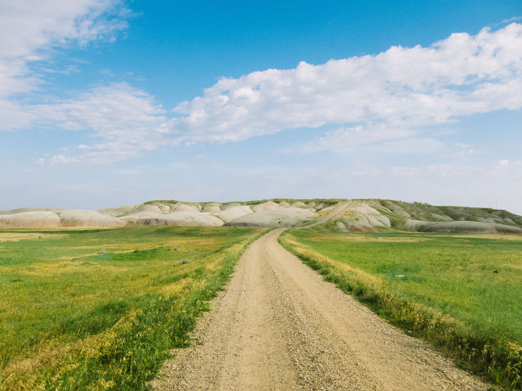Dirt road leading through green grassy fields toward low, rounded hills under a partly cloudy blue sky.