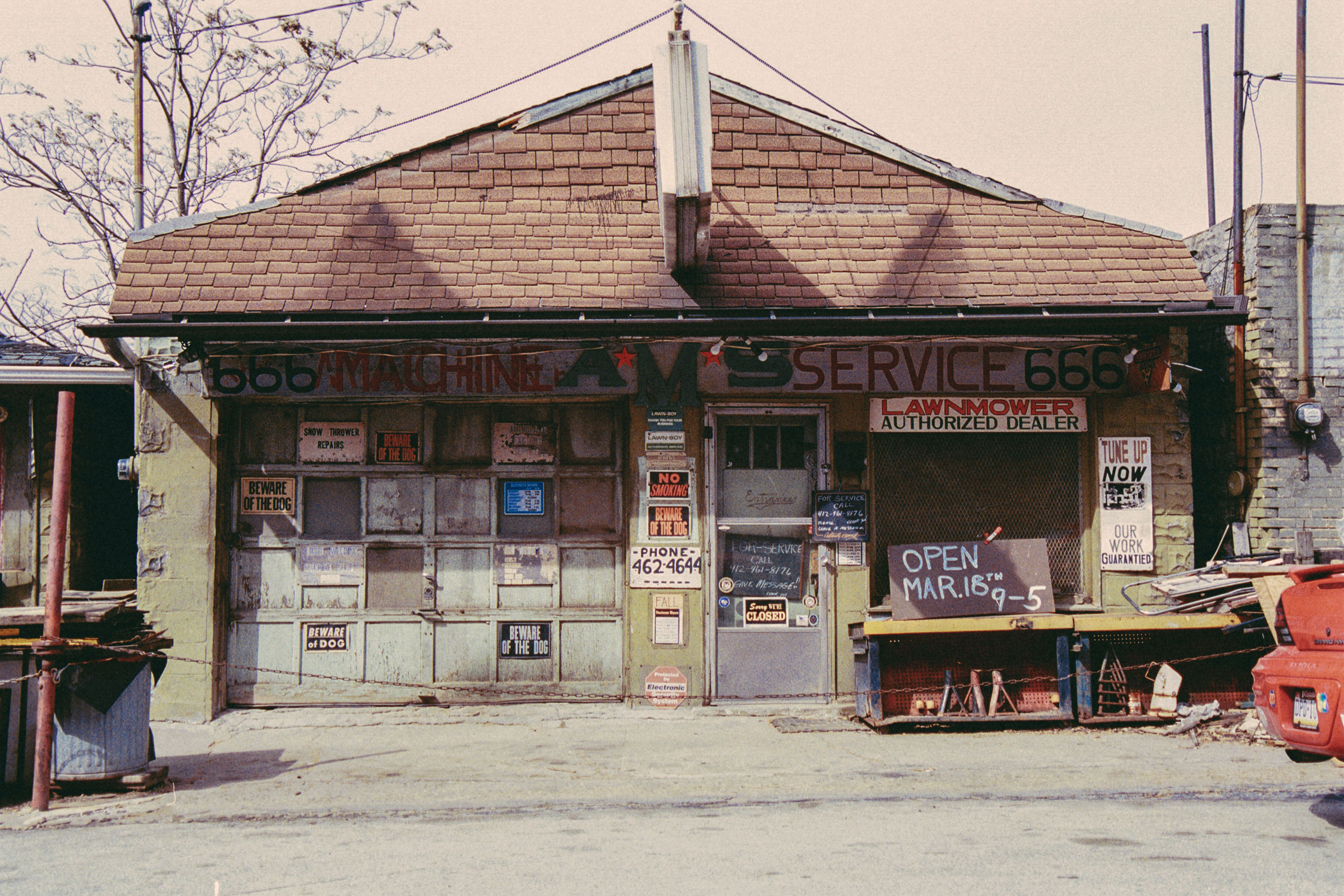 Old garage building with a brown shingled roof and faded signage reading 'AMS Machine Service 666'; various signs on the garage door and entrance door include 'Beware of the Dog,' 'No Smoking,' 'Lawnmower Authorized Dealer,' and 'Open Mar. 18th 9-5.' A red pickup truck is partially visible on the right side.
