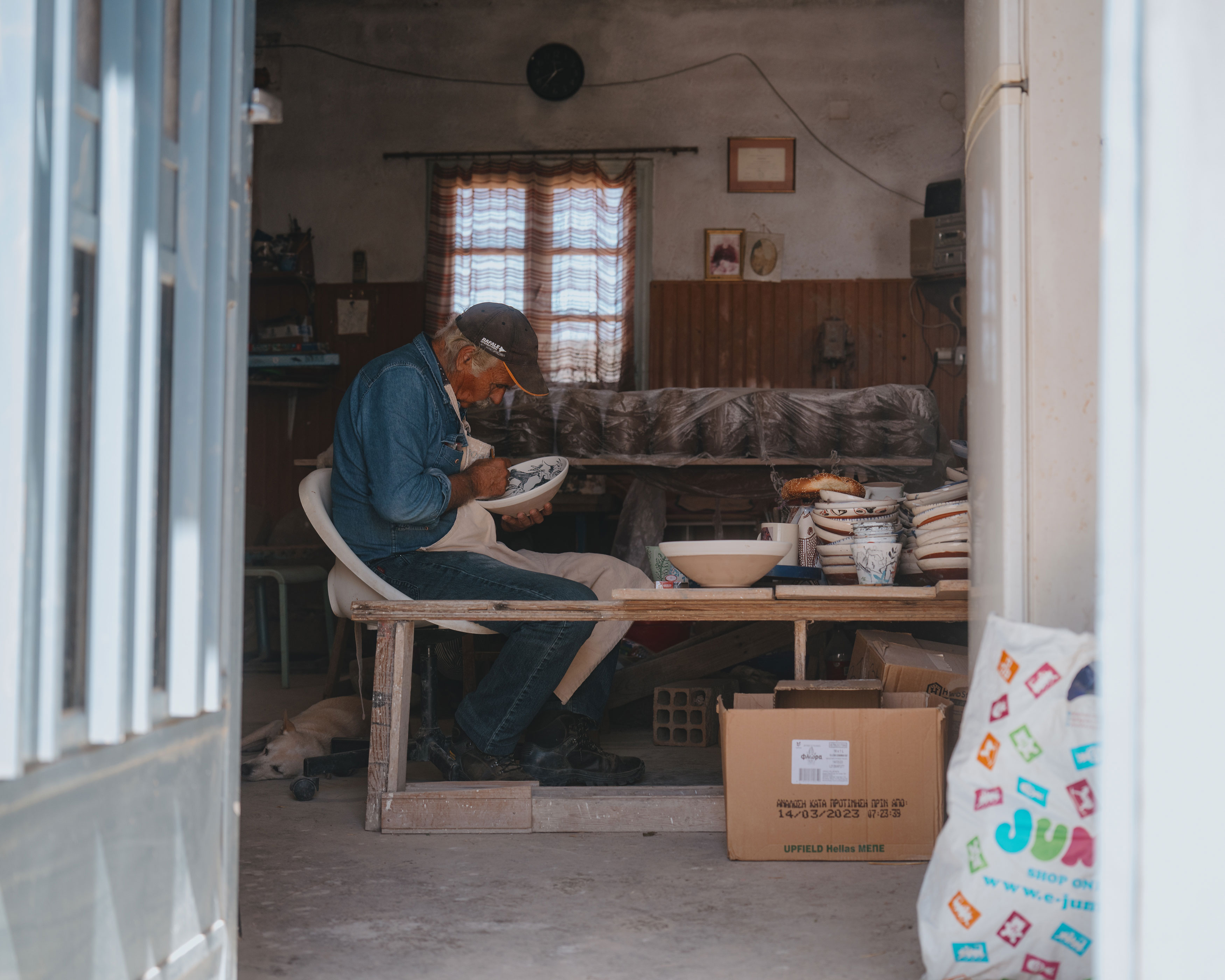 An elderly man wearing a denim shirt and apron sits on a white chair inside a workshop, carefully painting a ceramic bowl with a brush, surrounded by stacked pottery and boxes.