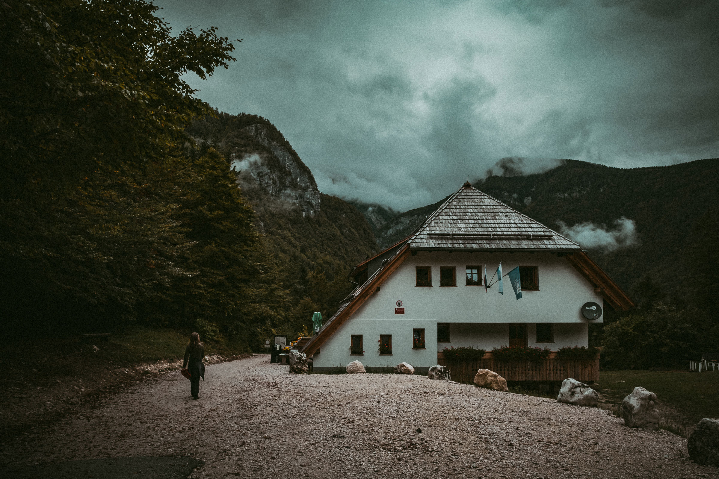 Person walking on a gravel path beside a white house with a steep roof, set against forested mountains under a cloudy, moody sky.