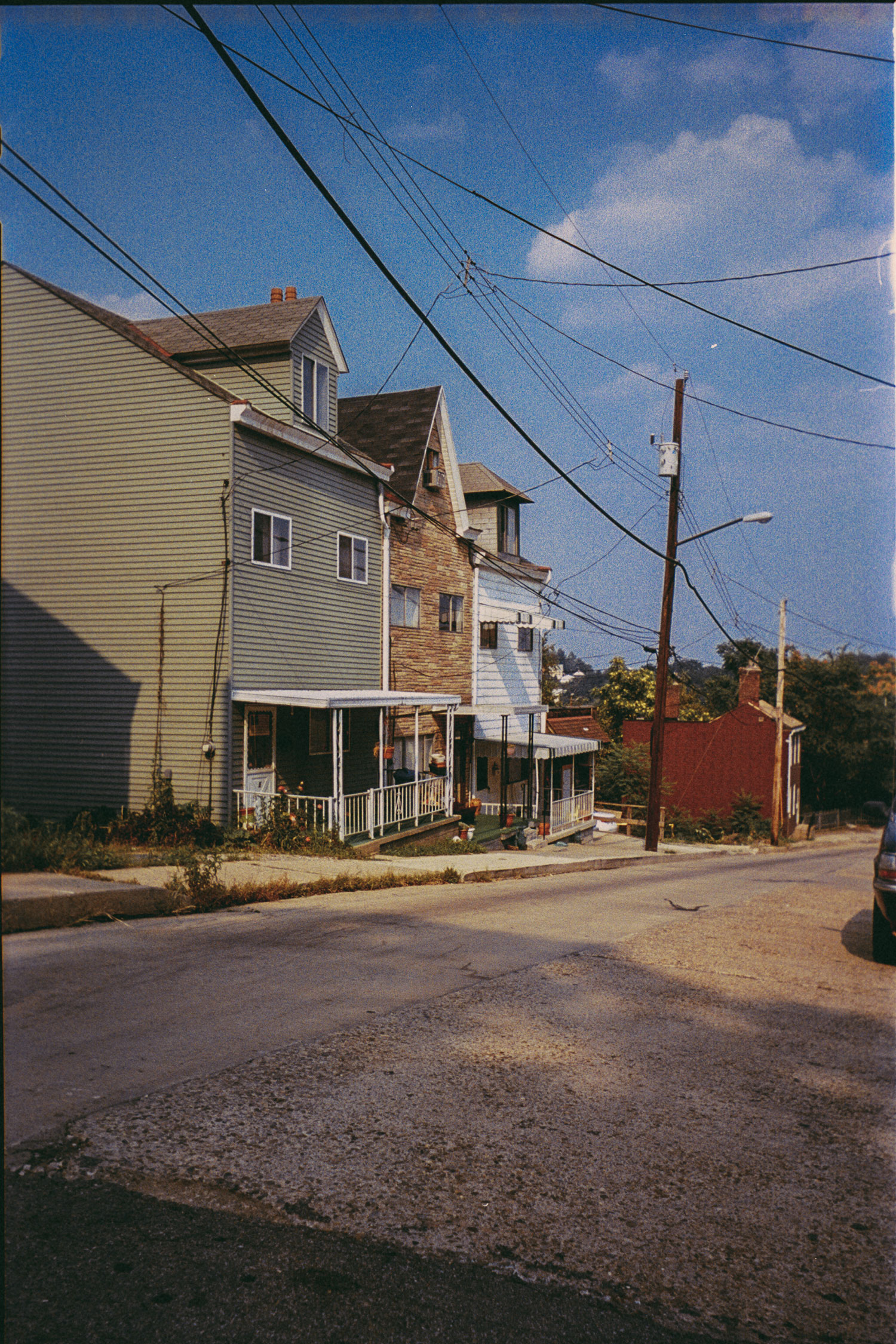 Street view of a quiet residential neighborhood with older two- and three-story houses featuring porches, utility poles with overhead power lines, a partly cloudy blue sky, and a parked car on the right side of the paved road.