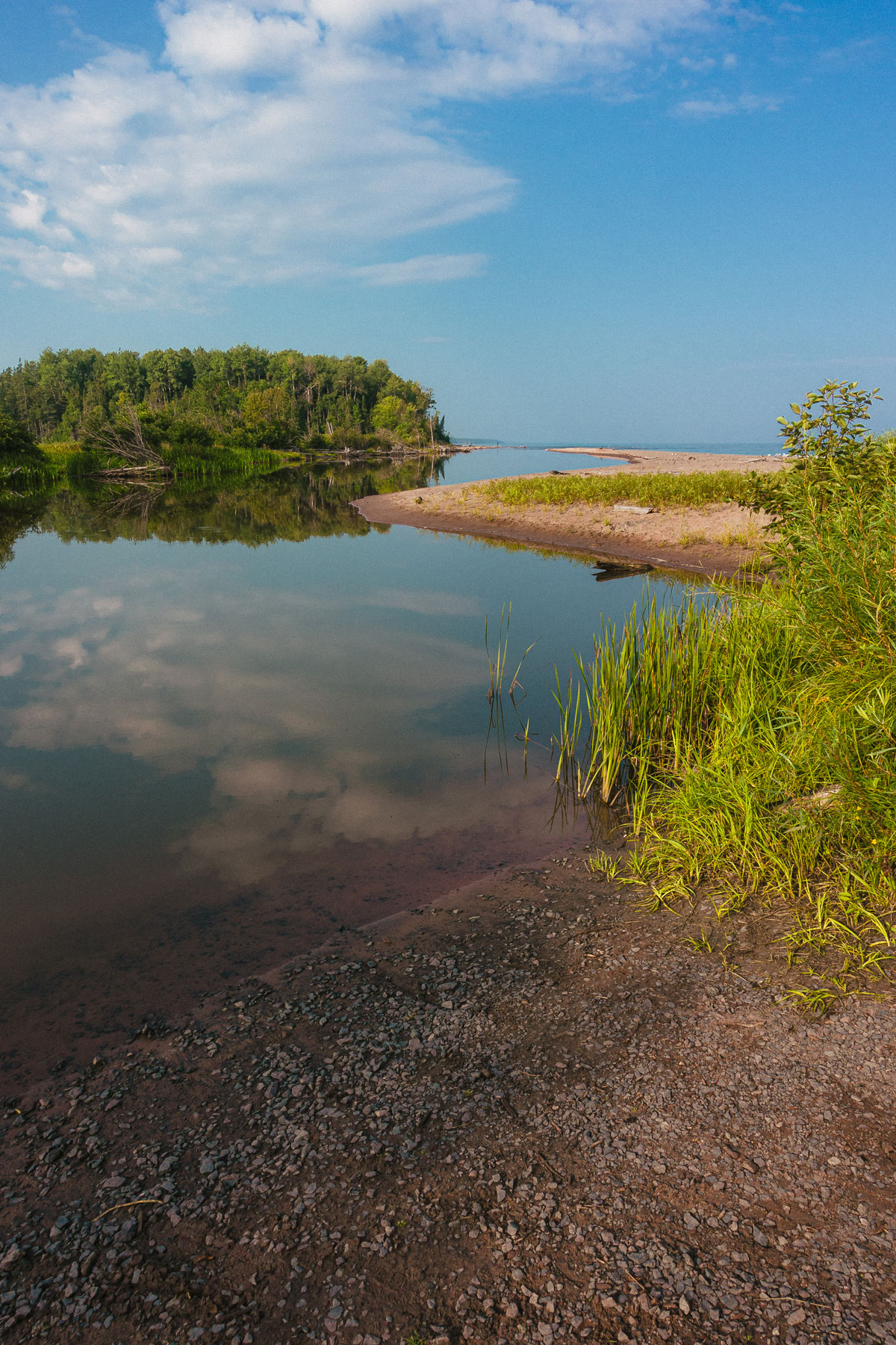 Calm river or lake shore with clear reflections of clouds and sky, bordered by green grass and trees, under a bright blue sky.