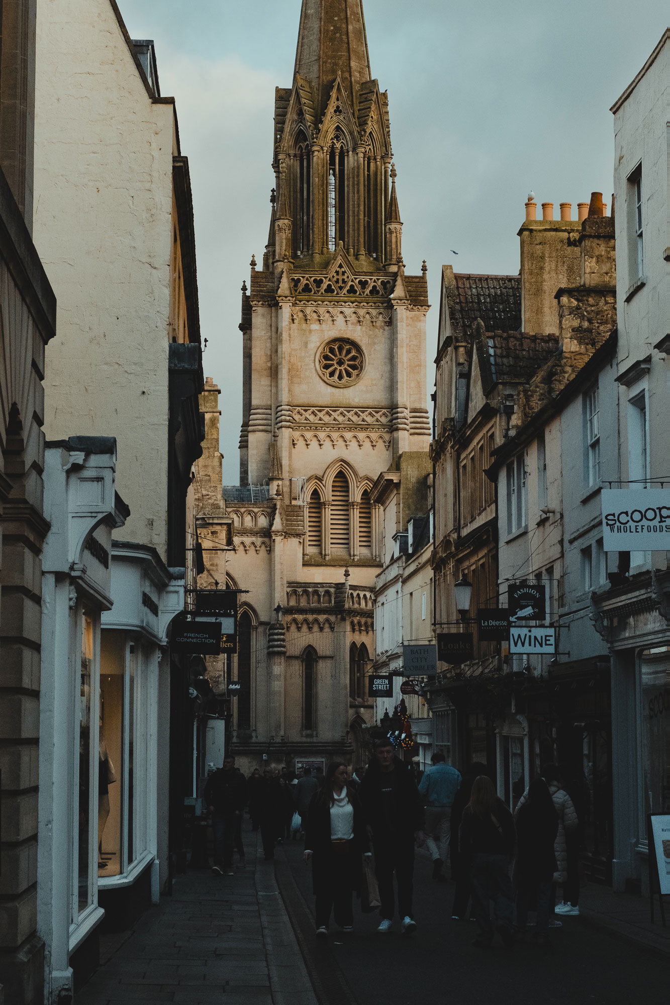 Narrow street with pedestrians walking near shops and a tall Gothic-style church tower with detailed stonework against a cloudy sky.