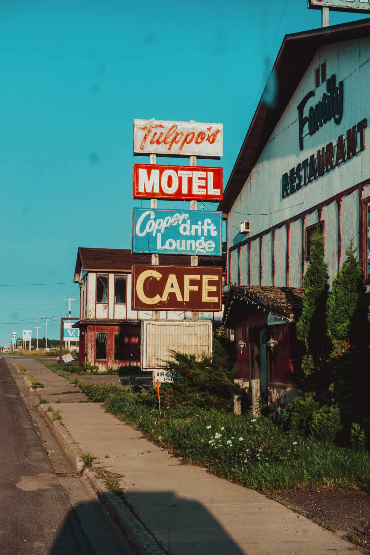 Vintage roadside signs reading 'Tulppo's Motel,' 'Copper drift Lounge,' and 'Cafe' mounted on poles next to a sidewalk with buildings including a 'Family Restaurant' under a clear blue sky.