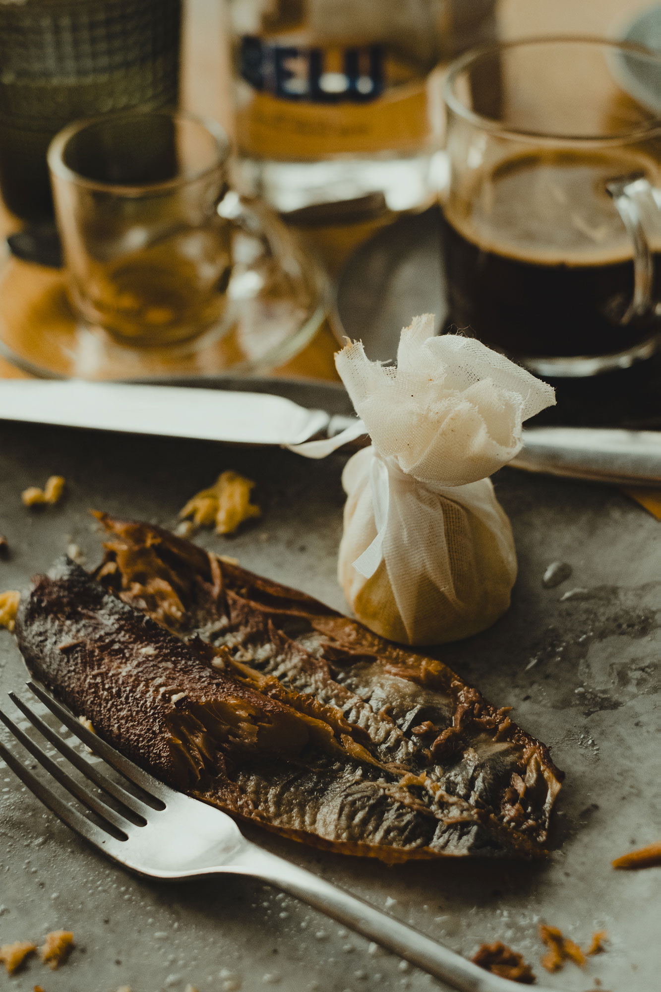 A cooked fish fillet on a plate with a lemon wrapped in cheesecloth nearby, a fork and knife rest on the plate, with blurred cups and a glass bottle in the background.