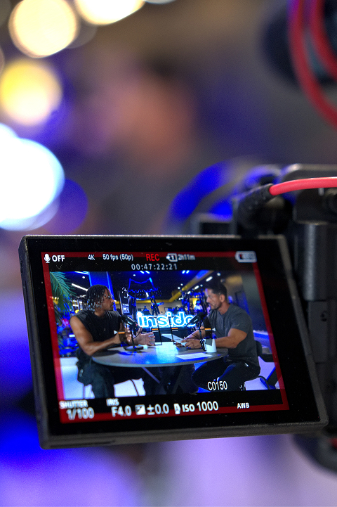 Camera screen showing two men seated at a round table with microphones, recording a podcast or interview in a brightly lit studio with the word 'inside' visible in the background.