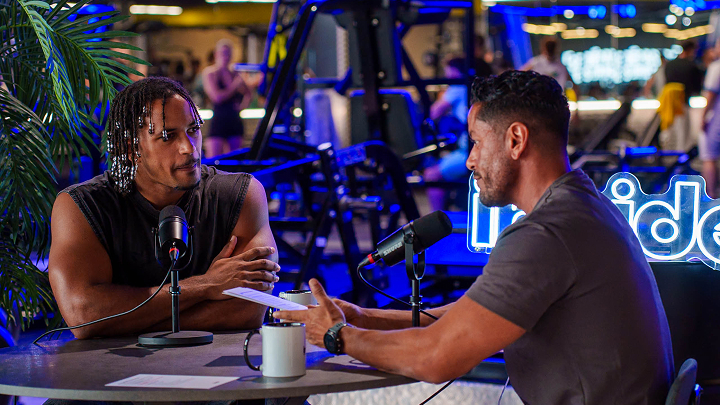 Two men seated at a table with microphones having a podcast conversation in a gym setting with workout equipment visible in the background.