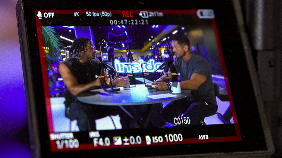 Camera screen showing two men sitting at a round table with microphones, engaged in a recorded conversation or podcast with a neon sign reading 'Inside' in the background.