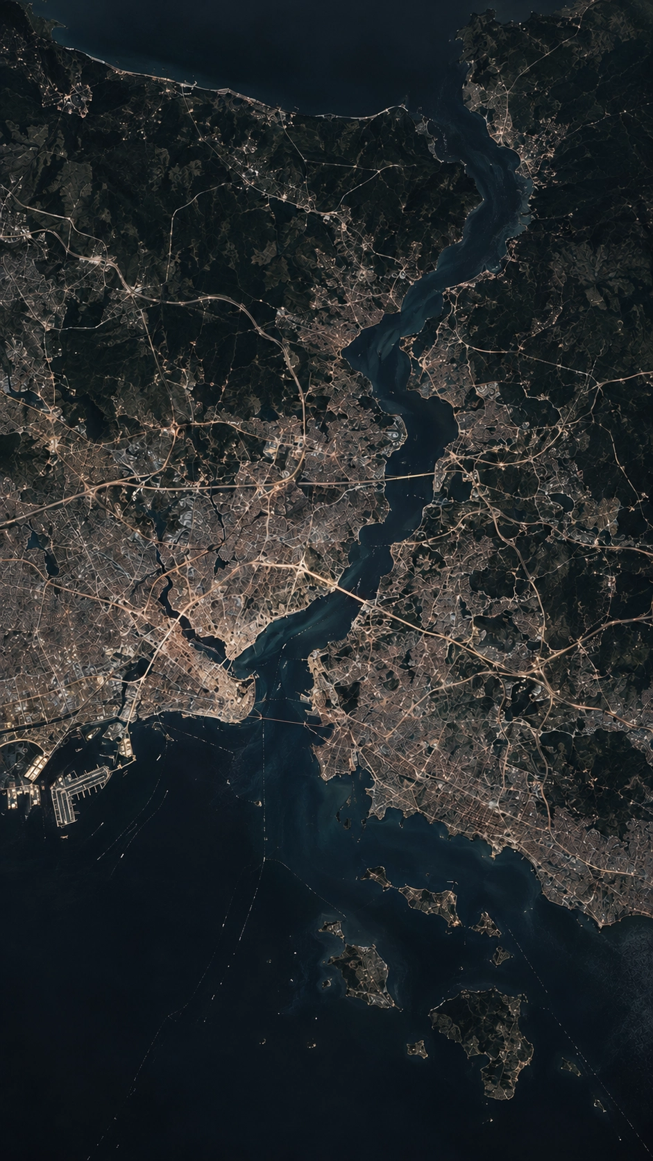 Nighttime satellite view of a large city divided by a strait with numerous illuminated roads, bridges, and islands along the water.