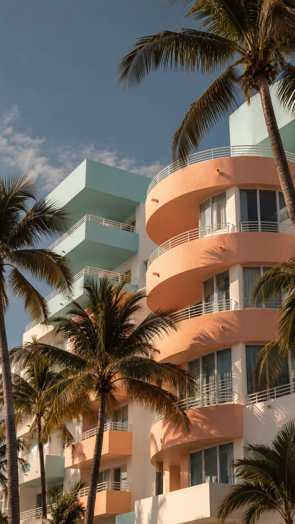 Sunlit building with pastel peach and teal balconies framed by tall palm trees under a partly cloudy blue sky.
