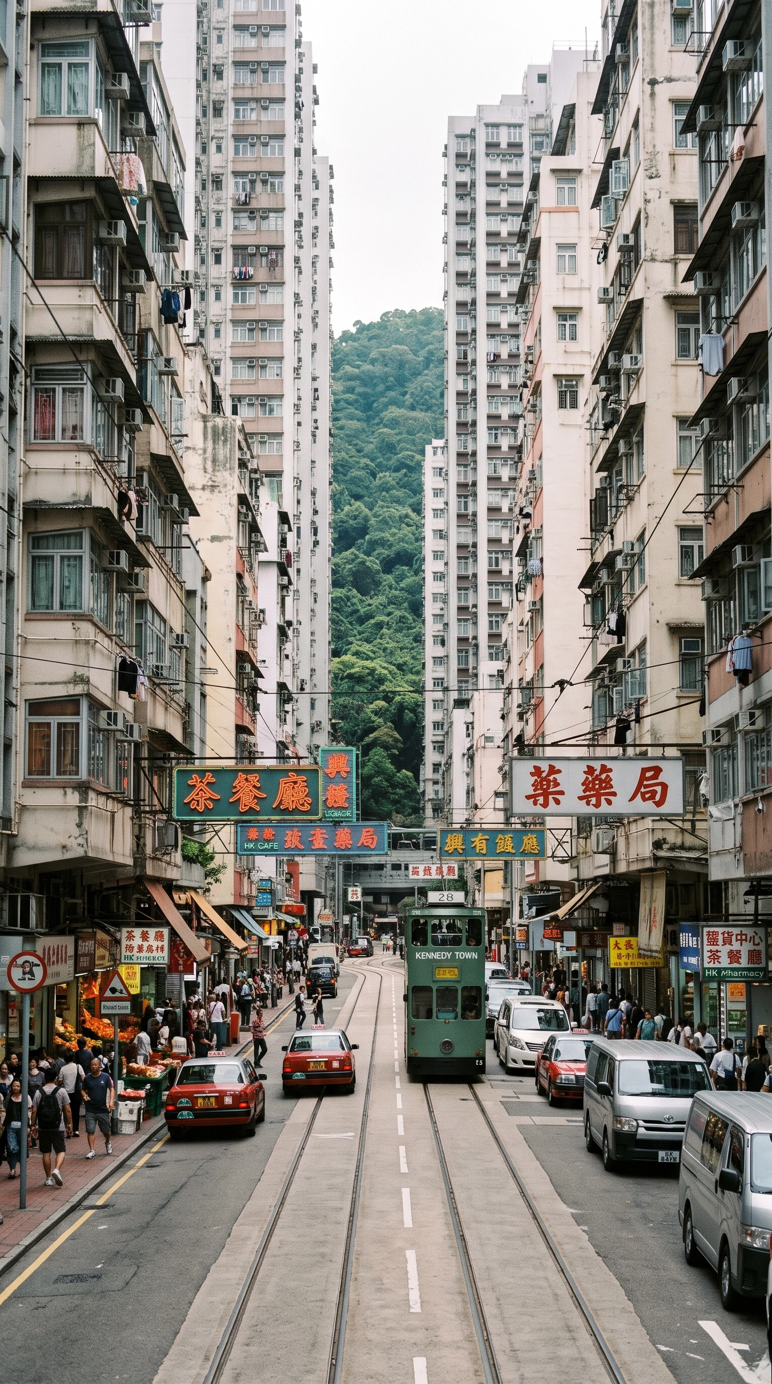 Urban street in Hong Kong with tall residential buildings on both sides, a green double-decker tram labeled Kennedy Town traveling on central tram tracks, parked red taxis and cars along the road, many pedestrians walking on sidewalks, and green forested hills visible in the background, under an overcast sky.