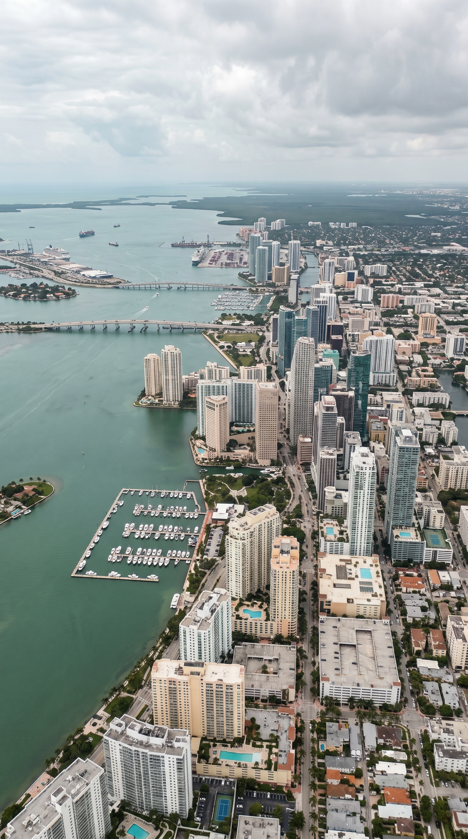 Aerial view of a coastal city with tall skyscrapers, residential buildings, swimming pools, and a marina filled with boats, under a cloudy sky.