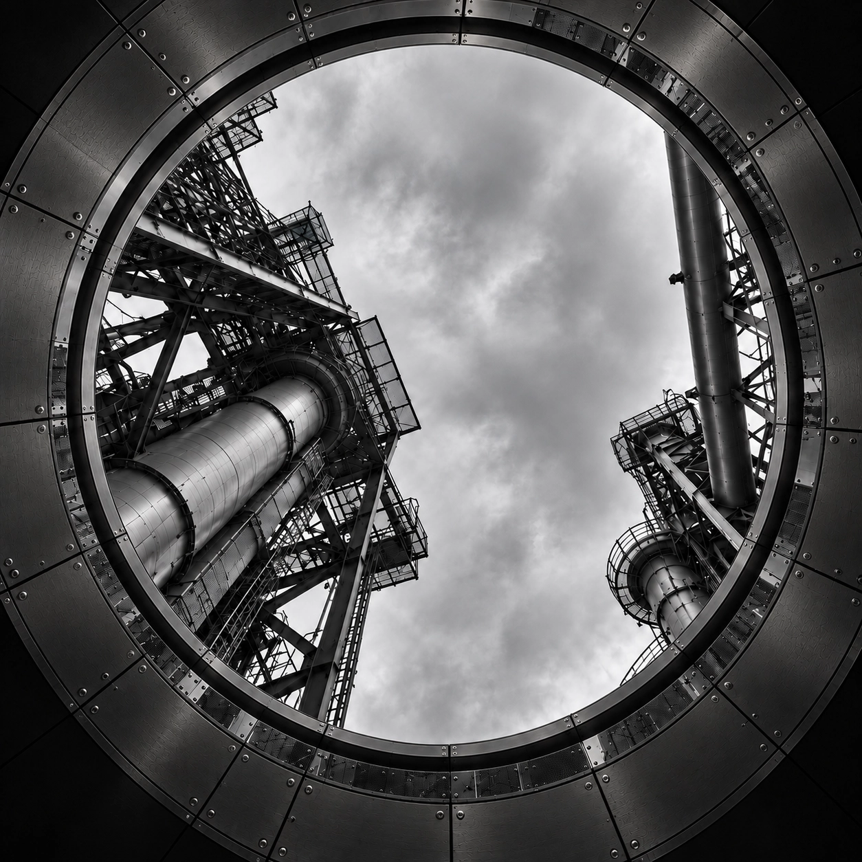 Looking upward through a circular metal frame at two tall industrial metal structures with ladders and platforms against a cloudy sky in black and white.