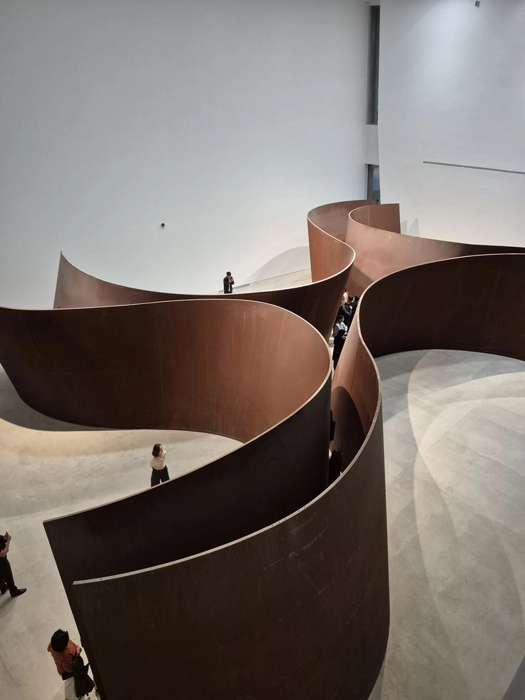 Large abstract indoor sculpture with tall, curving, brown metal walls forming winding paths, with visitors walking and standing inside a spacious gallery with white walls and concrete floor.