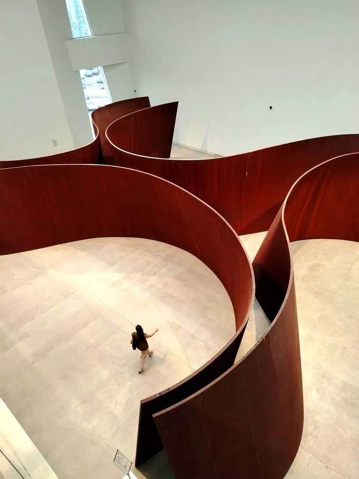 Person walking inside a large indoor maze made of tall curved rust-colored metal walls on a light floor, with white walls and tall window in the background.