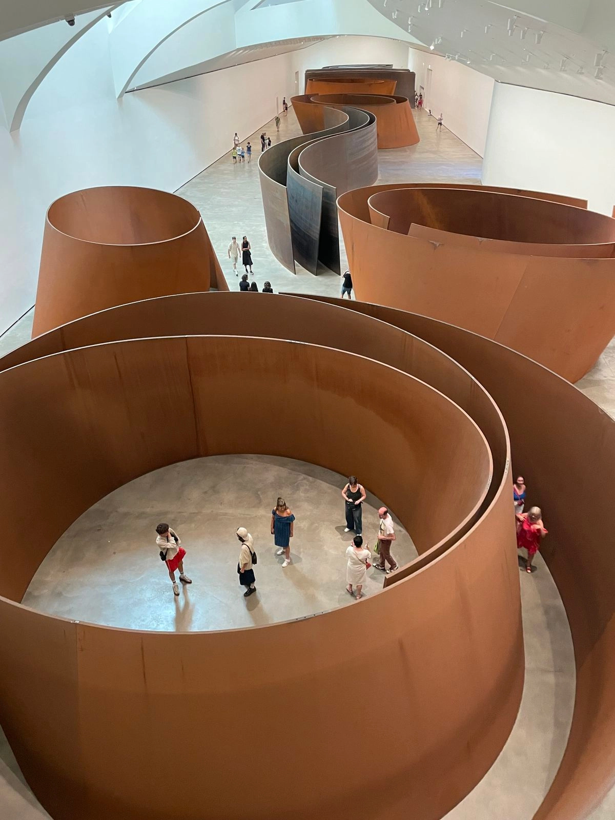Large indoor art installation featuring tall, curved rust-colored metal walls in a spiral and wave pattern with visitors walking inside and around them in a spacious gallery with white walls and ceiling.