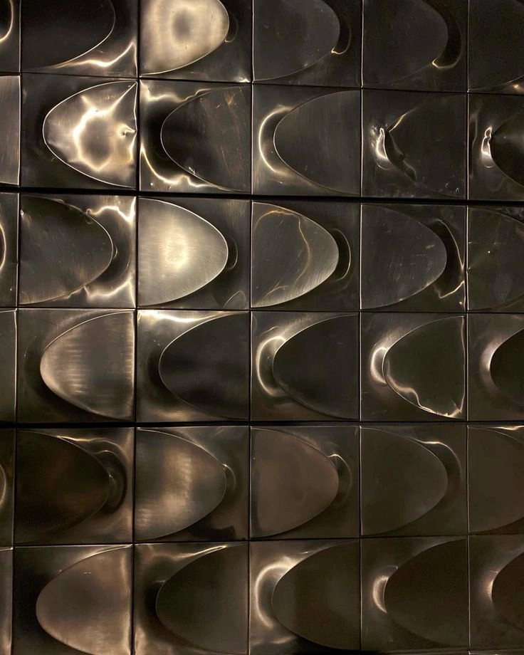 Close-up of a wall covered with metallic tiles featuring a repetitive pattern of curved, embossed shapes reflecting light.