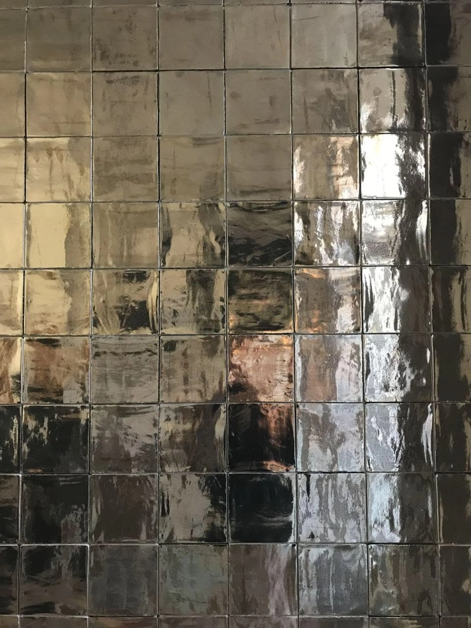 Wall covered with glossy, reflective square tiles in varying shades of brown, bronze, and black, creating a mosaic with light reflections.