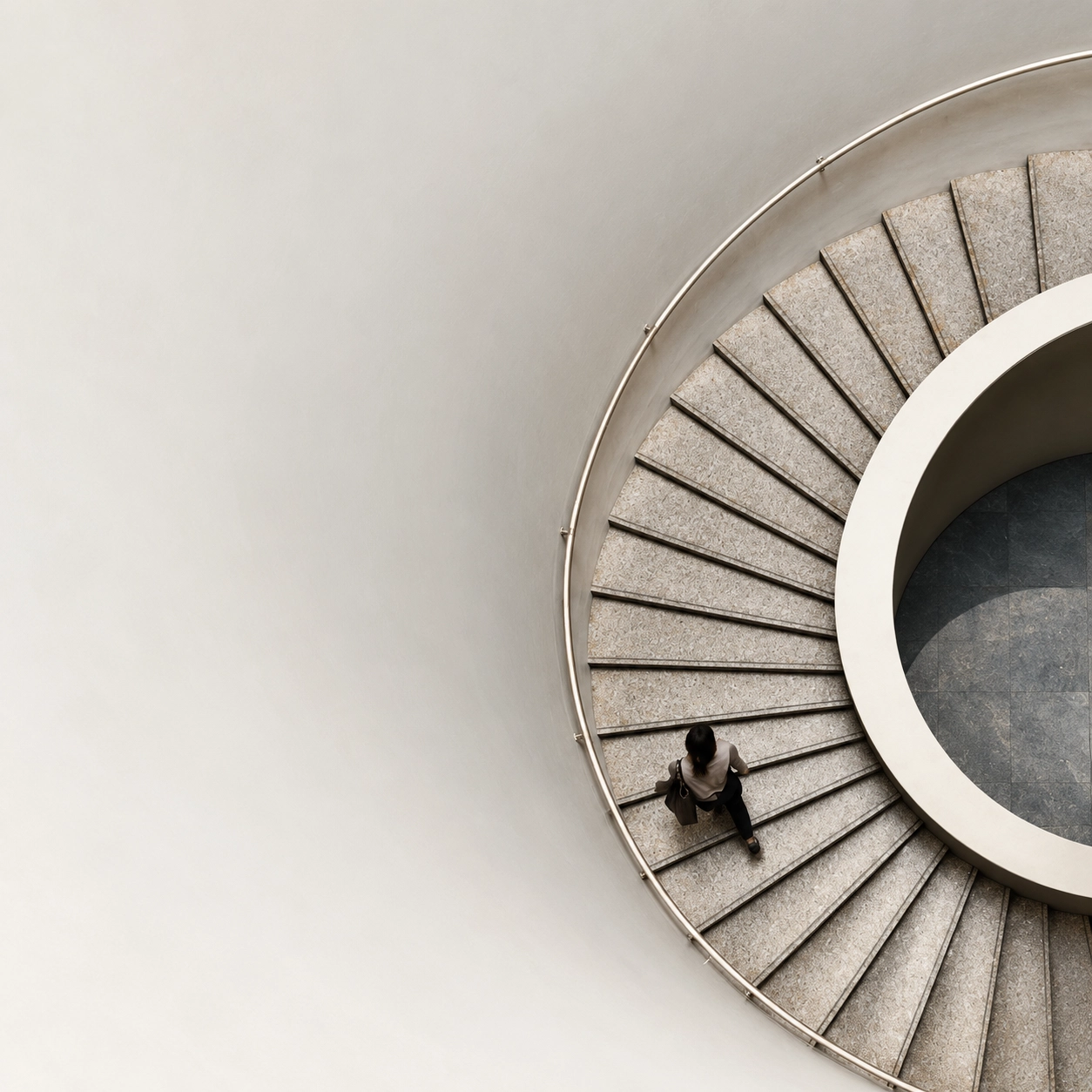 Top-down view of a person walking up a curved stone spiral staircase with a beige wall and simple metal railing.
