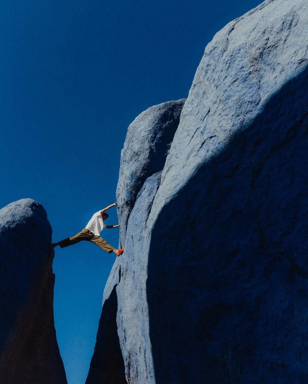 Person wearing a white shirt, brown pants, and red shoes stretching between two large blue-gray rocks under a clear blue sky.
