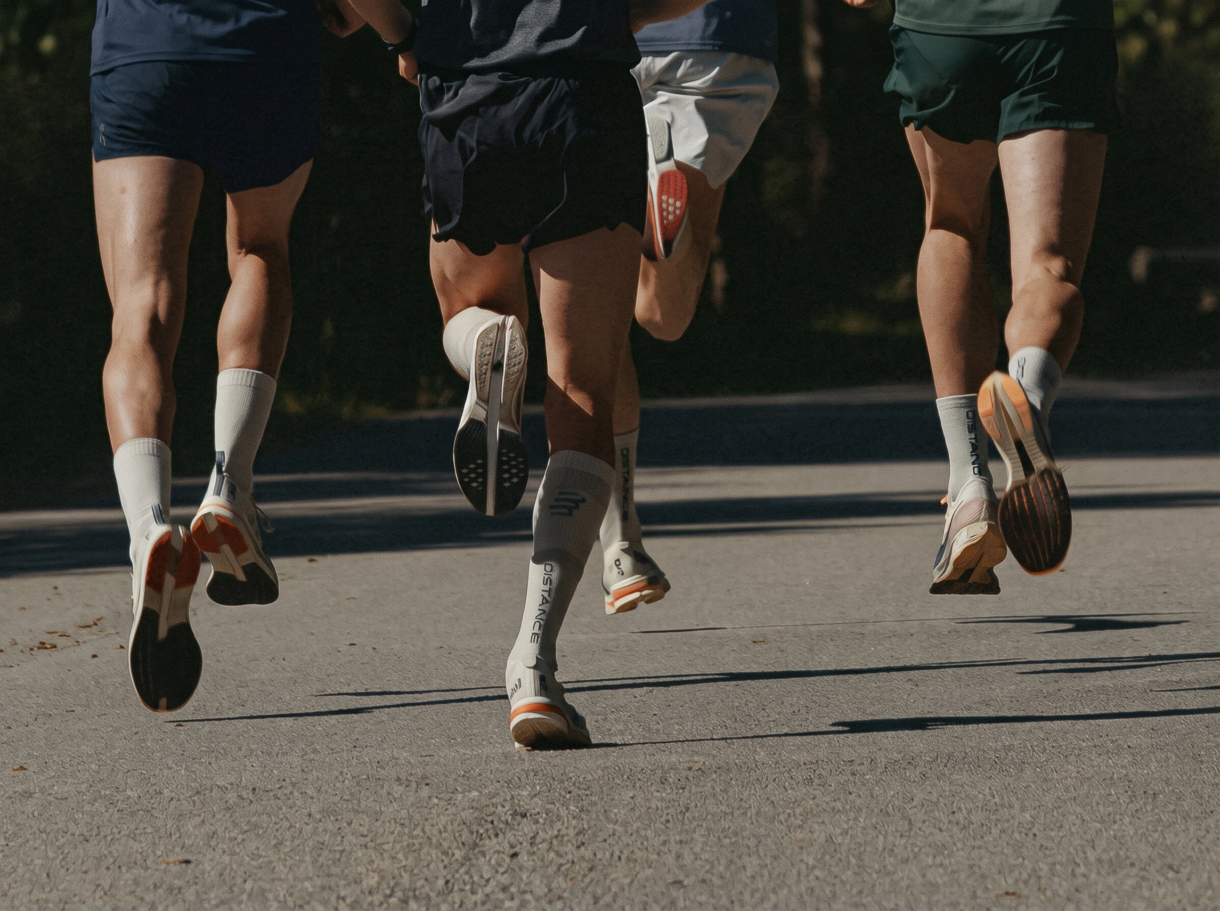 Close-up of four people running on a paved road, focusing on their legs and running shoes, all wearing white socks and athletic shorts.