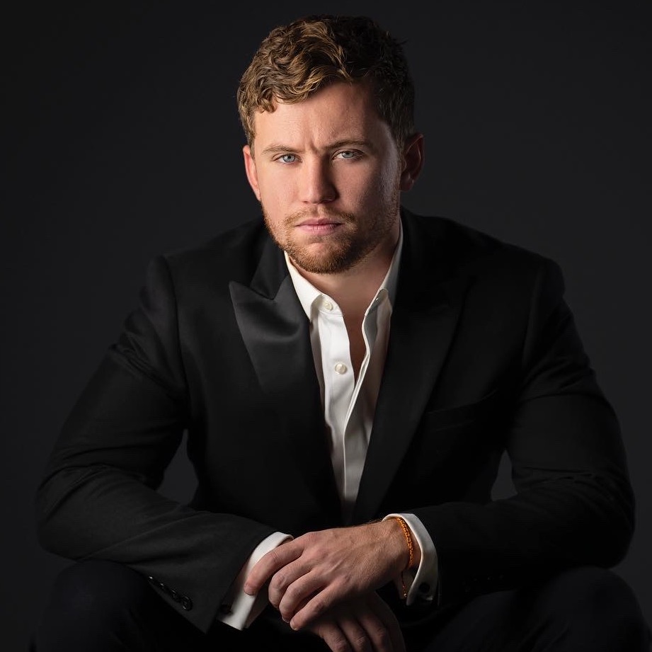 Professional headshot of a man in a black tuxedo jacket seated against a dark background