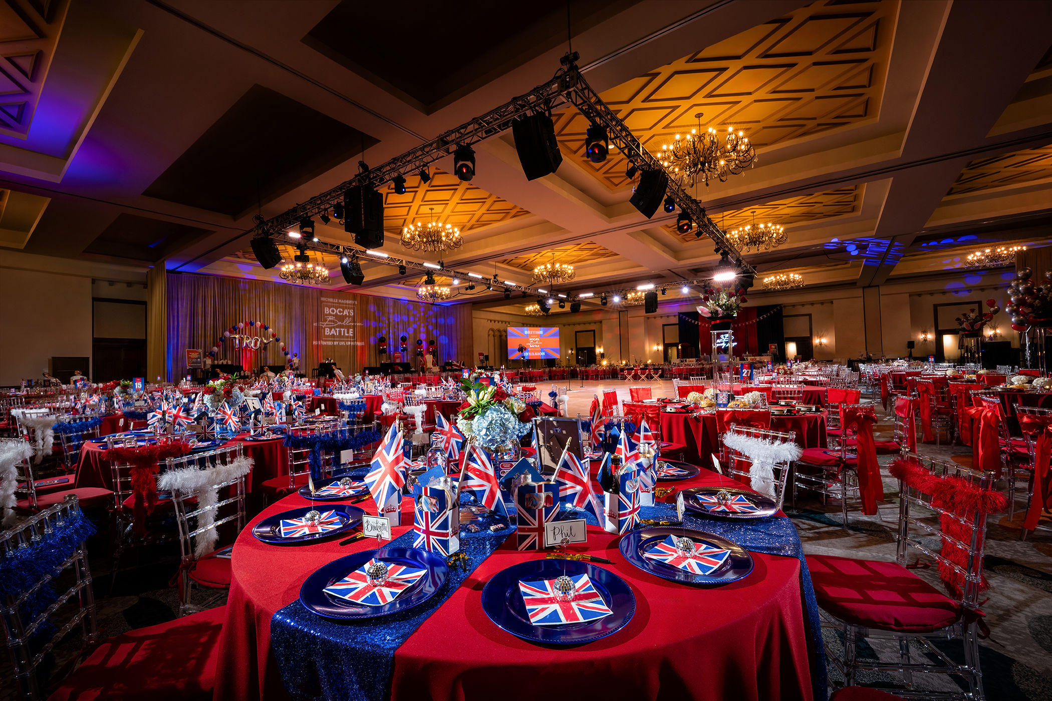 Ballroom set for a British-themed gala with Union Jack place settings, red linens and stage lighting