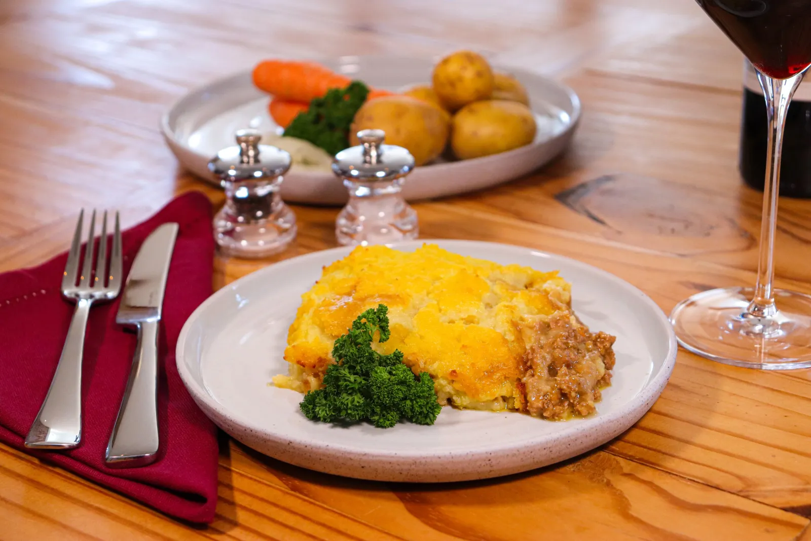 A mash-topped pie with parsley on a wooden table, with carrots, potatoes and a glass of red wine.