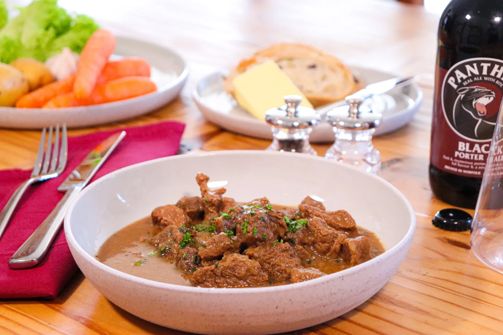 Beef casserole in a stoneware bowl beside a bottle of Panther Black Porter, with bread, butter and vegetables behind.