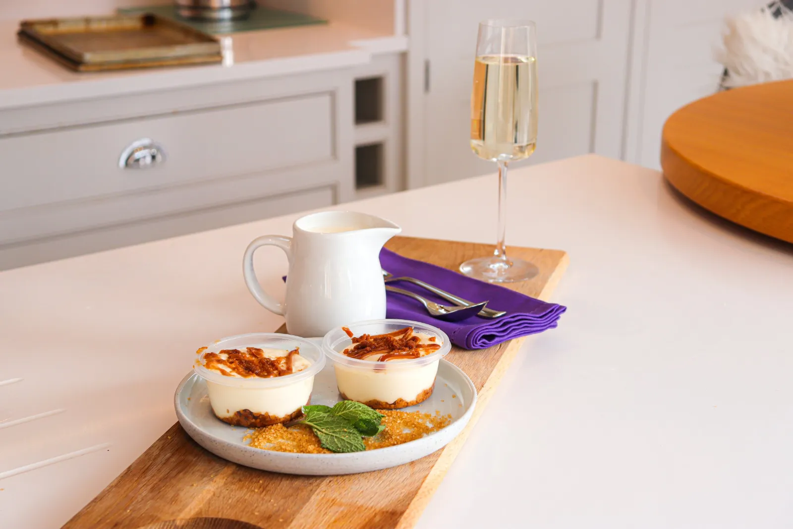 Two individual cheesecakes with a caramel drizzle, a jug of cream and a glass of sparkling wine in a country kitchen.