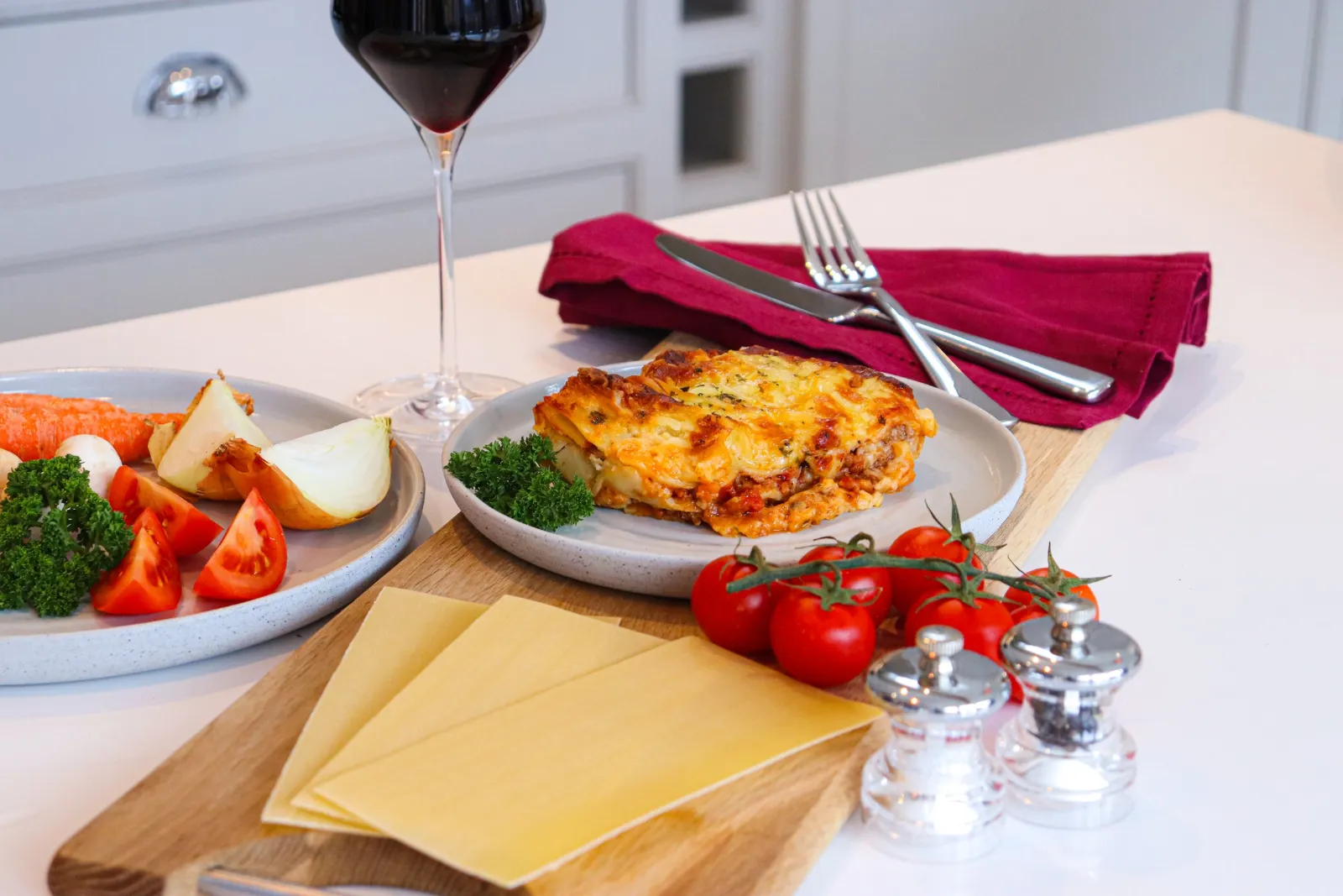 A portion of lasagne on an oak board with fresh pasta sheets, vine tomatoes and a glass of red wine.