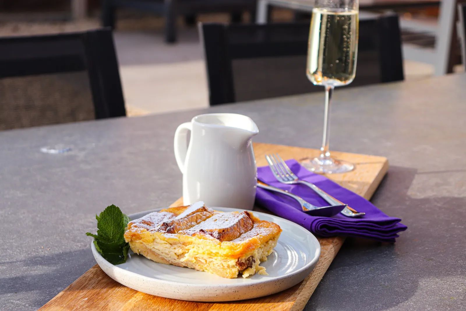 Bread and butter pudding with a jug of cream and a glass of sparkling wine, on an outdoor table in the sun.