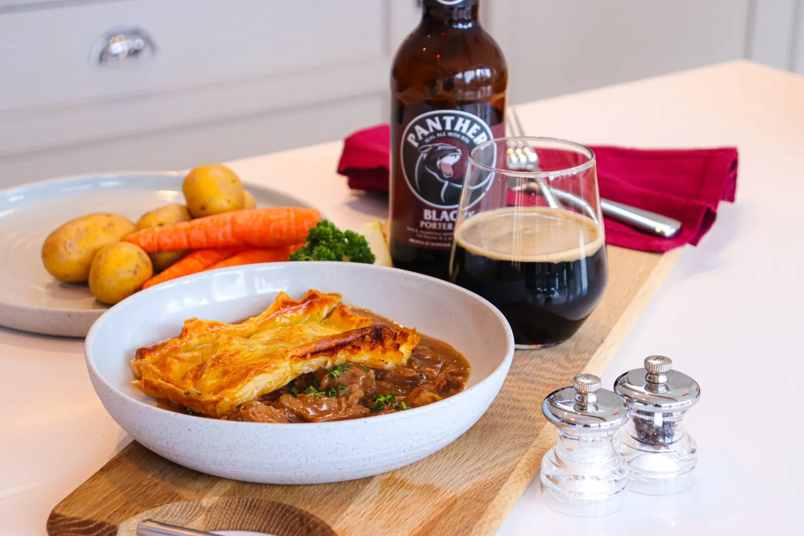 A steak and ale pie in a stoneware bowl beside a bottle and glass of Panther Black Porter, on an oak board in a country kitchen.
