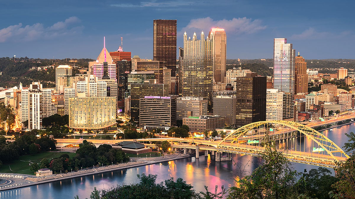 Dusk skyline with illuminated towers, a yellow steel bridge, rivers, and wooded hills.