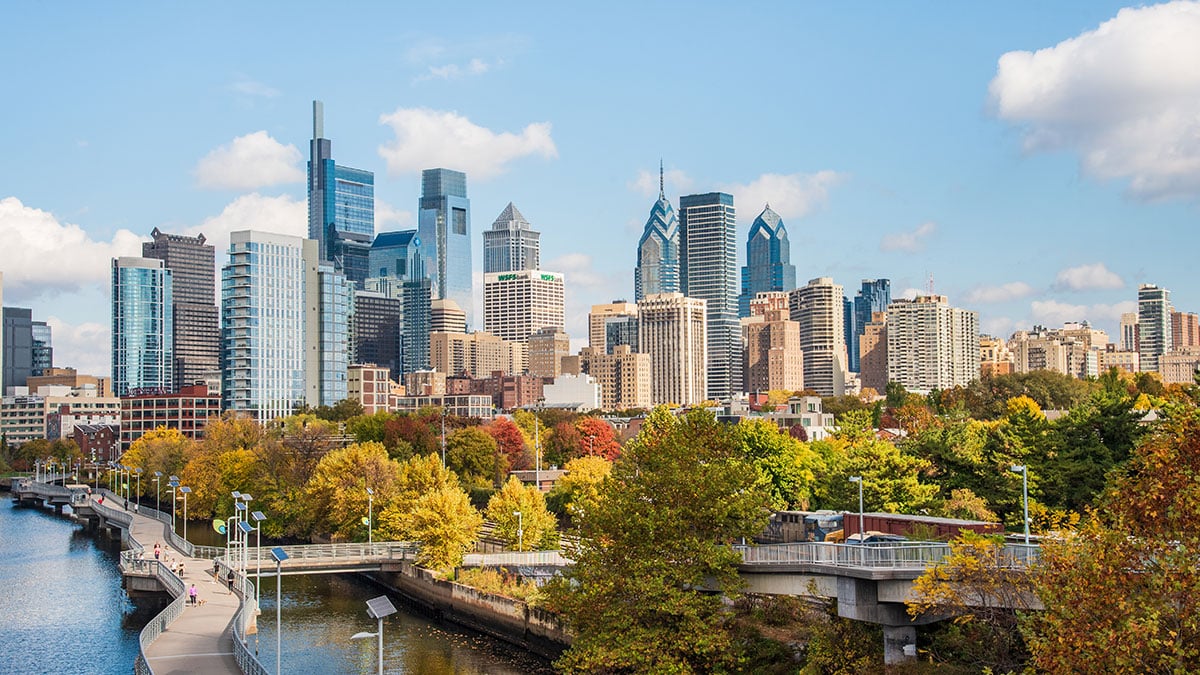 Autumn waterfront park with pedestrian bridges and trees in front of a dense modern city skyline.
