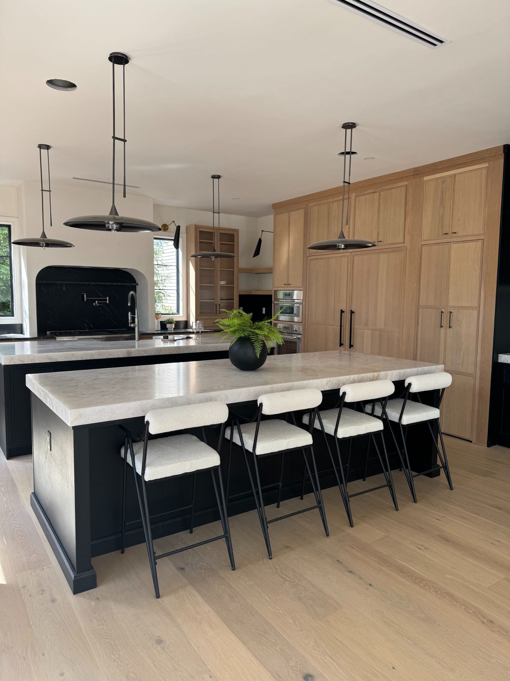 Light oak cabinets with a dark navy island, white counter stools and a pale stone counter in an open kitchen