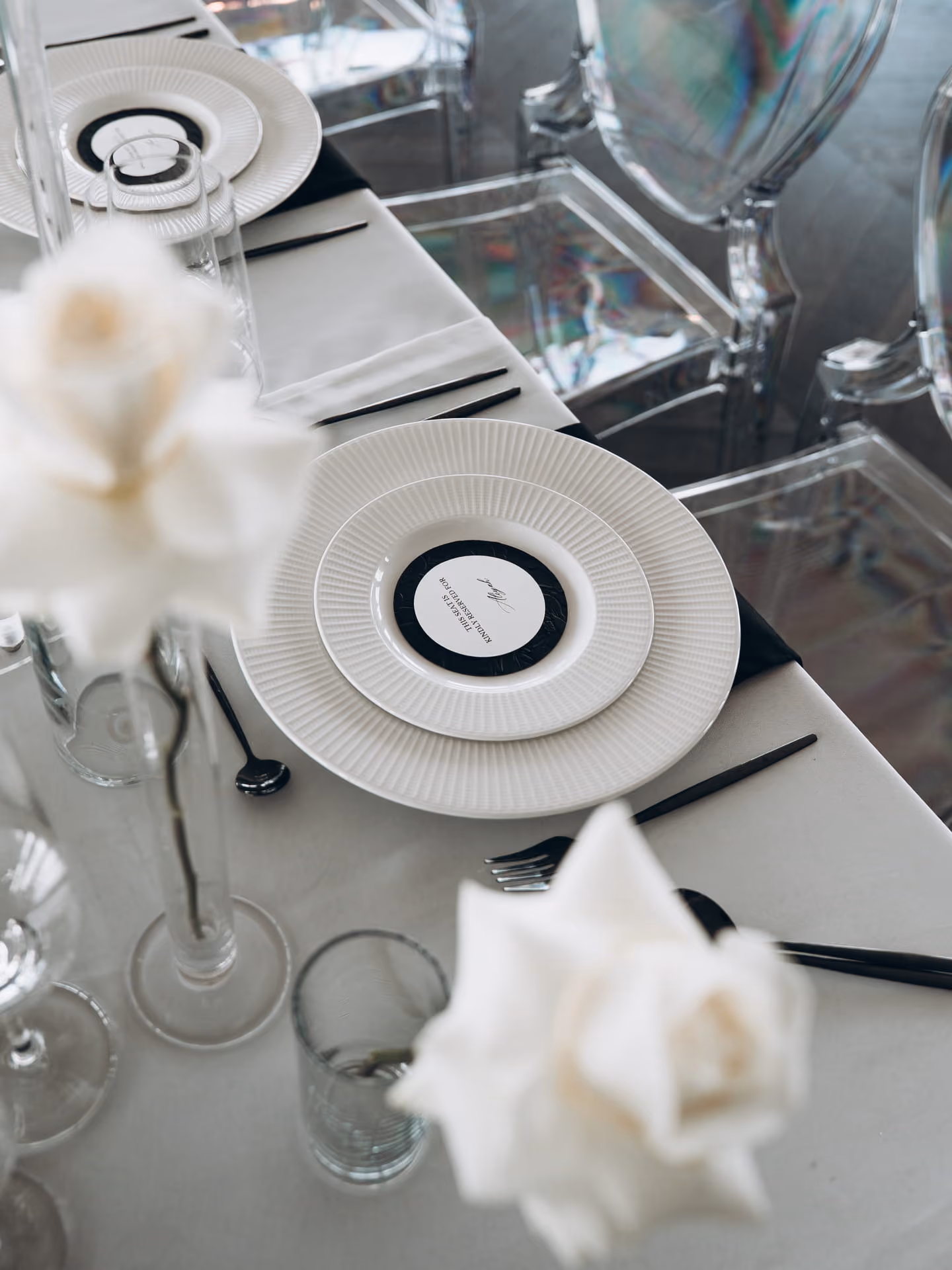 Eleganter gedeckter Tisch mit weißen Tellern, schwarzem Besteck und weißen Rosen in Glasvasen, durchsichtige Stühle im Hintergrund.
