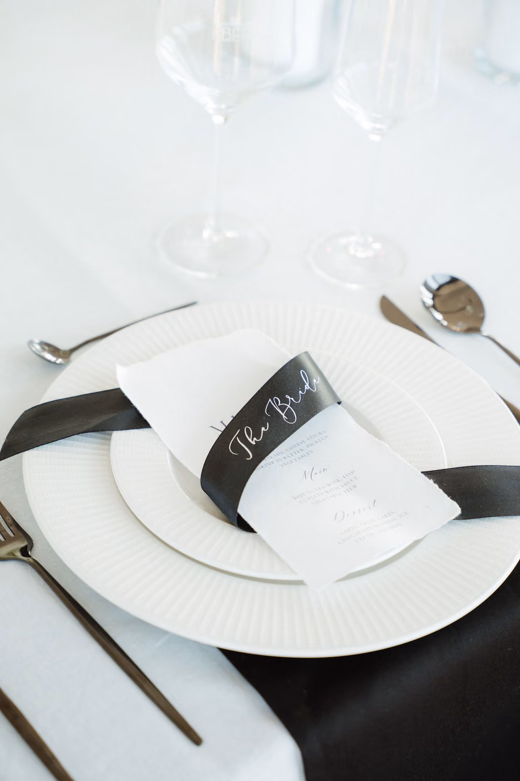 Eleganter gedeckter Hochzeitstisch mit weißem Geschirr, schwarzem Besteck und einem weißen Menü auf dem Teller, gehalten von einem schwarzen Band mit der Aufschrift „The Bride“.
