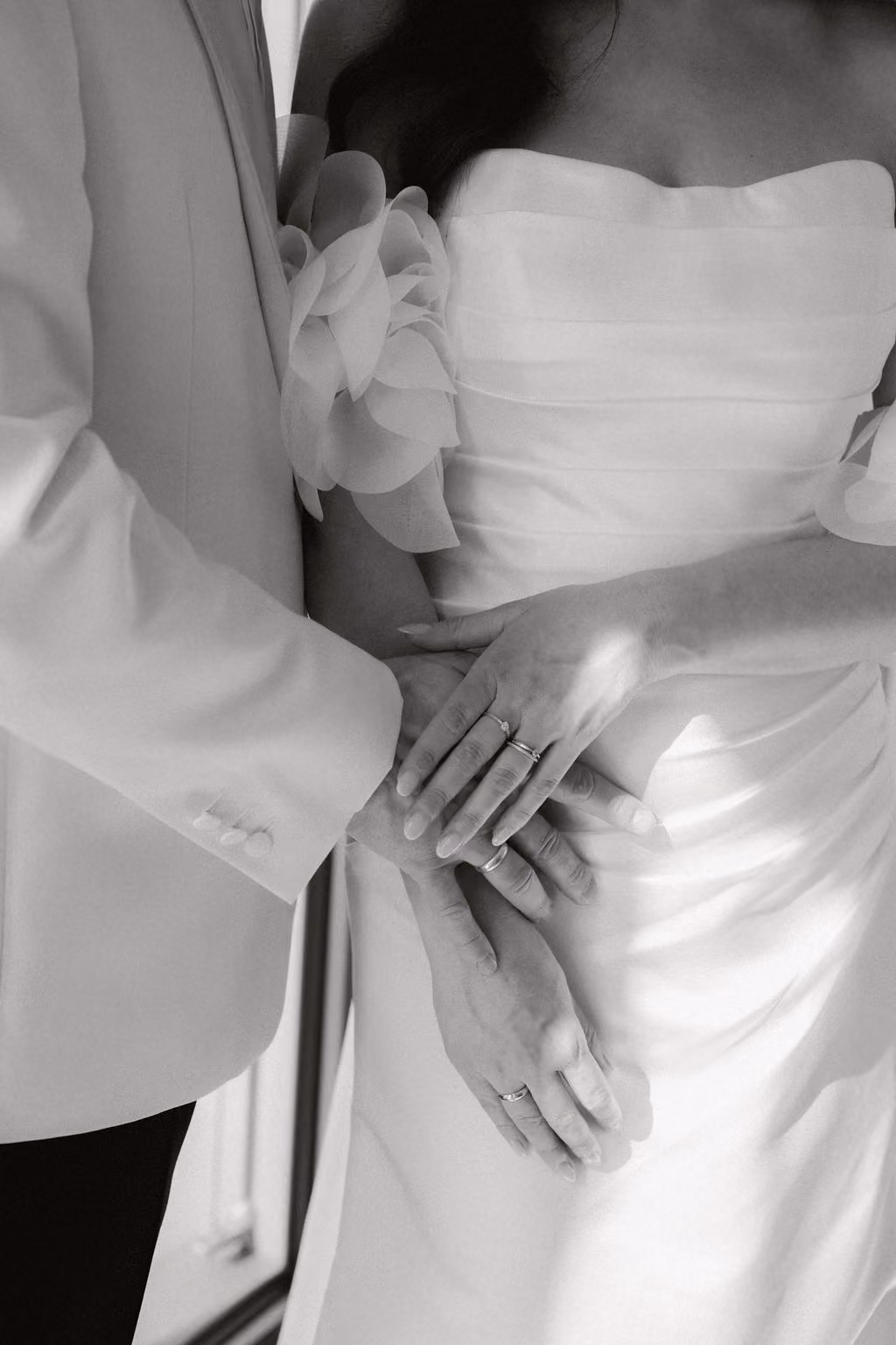 Schwarz-weißes Foto eines Brautpaares, das Hände mit Eheringen hält, wobei die Braut ein ärmelloses Kleid mit Blumenärmeln trägt und der Bräutigam einen hellen Anzug.
