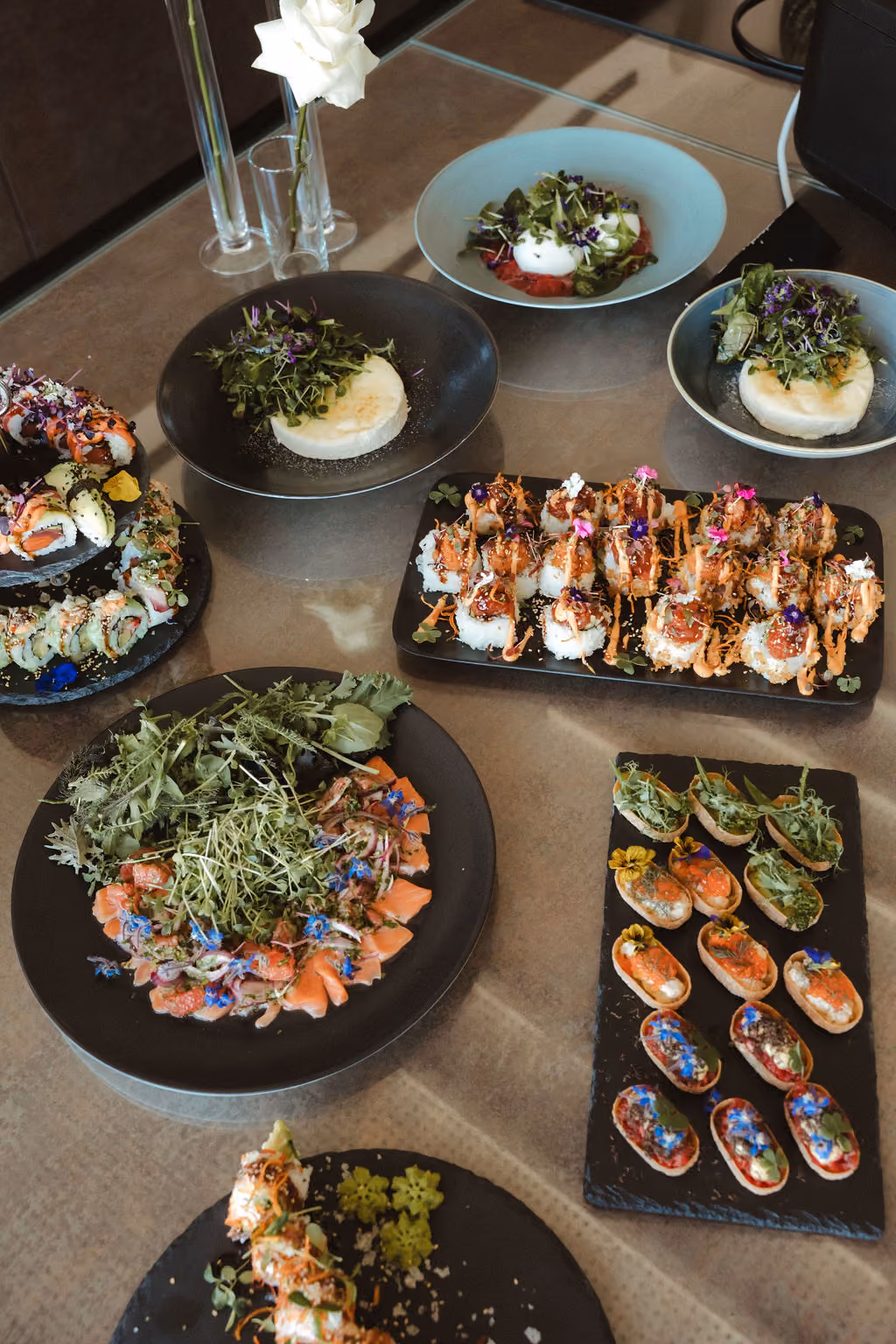 Vielfältige, kunstvoll angerichtete Speisen auf Tellern und Platten, darunter Sushi-Rollen, kleine Canapés, ein Teller mit Lachs und Salat und drei Teller mit grünen Salaten und dekorativen Blüten, auf einem Glas- oder Glastisch mit einer weißen Rose in Glasvasen.