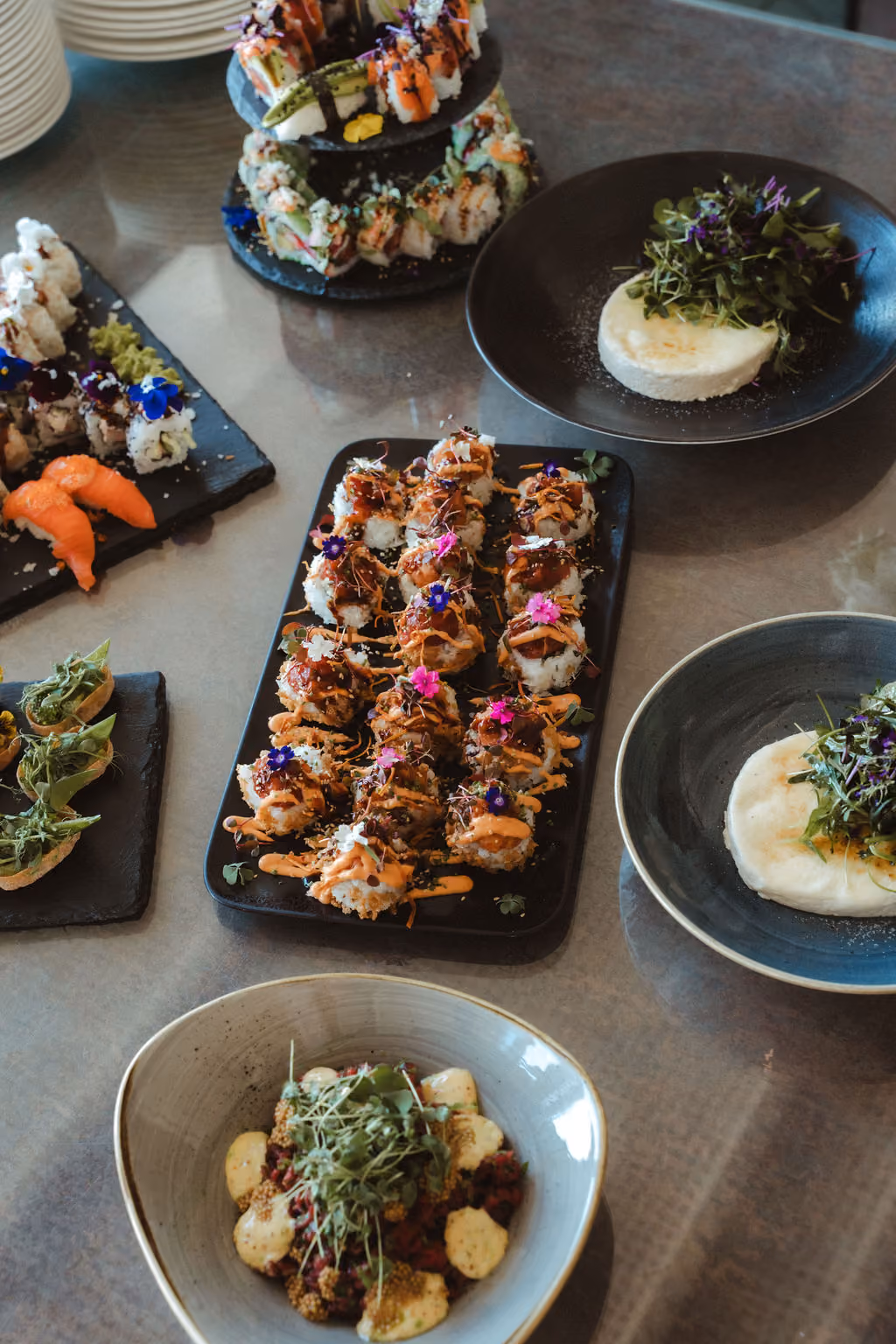 Mehrere Teller mit kunstvoll angerichtetem Sushi, darunter Rollen mit Saucen, Garnierungen und essbaren Blüten auf einem dunklen Tisch, begleitet von weiteren Schalen mit Salat und Kräutern.