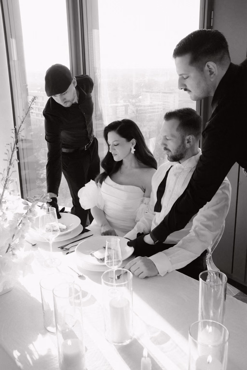 Schwarz-weiß-Foto eines Brautpaares bei einer Hochzeit, die an einem Tisch sitzen, während zwei männliche Kellner ihnen Speisen servieren. Die Braut trägt ein Schulterfreies Kleid mit Puffärmeln und der Bräutigam ein weißes Hemd mit Fliege. Der Tisch ist mit Kerzen in Glasbehältern, Weingläsern und Blumen dekoriert. Große Fenster im Hintergrund zeigen eine Stadtsicht.