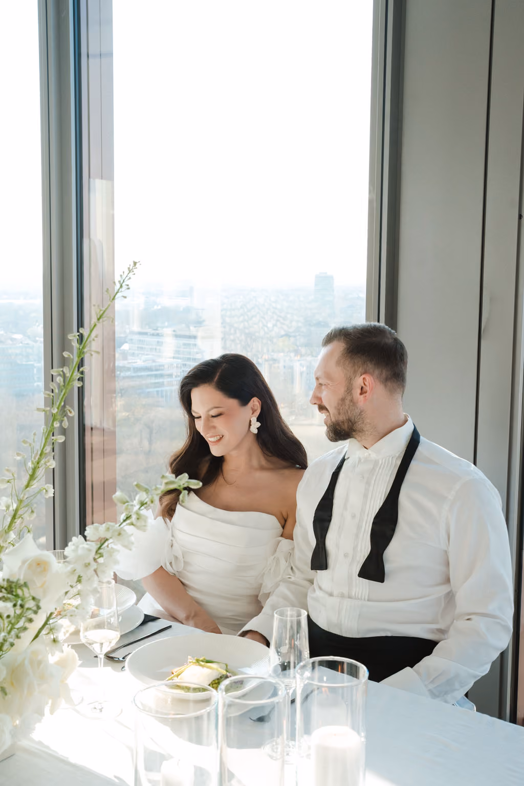 Ein Brautpaar sitzt an einem festlich gedeckten Tisch vor einem großen Fenster mit Blick auf die Stadt. Die Braut trägt ein schulterfreies weißes Hochzeitskleid mit Puffärmeln und auffälligen Ohrringen, sie lächelt und schaut nach unten. Der Bräutigam trägt ein weißes Hemd mit Fliege, die offen um den Hals hängt, und schaut sie lächelnd an. Auf dem Tisch stehen Gläser, Kerzen und ein Teller mit Essen. Weiße Blumen dekorieren den Tisch.