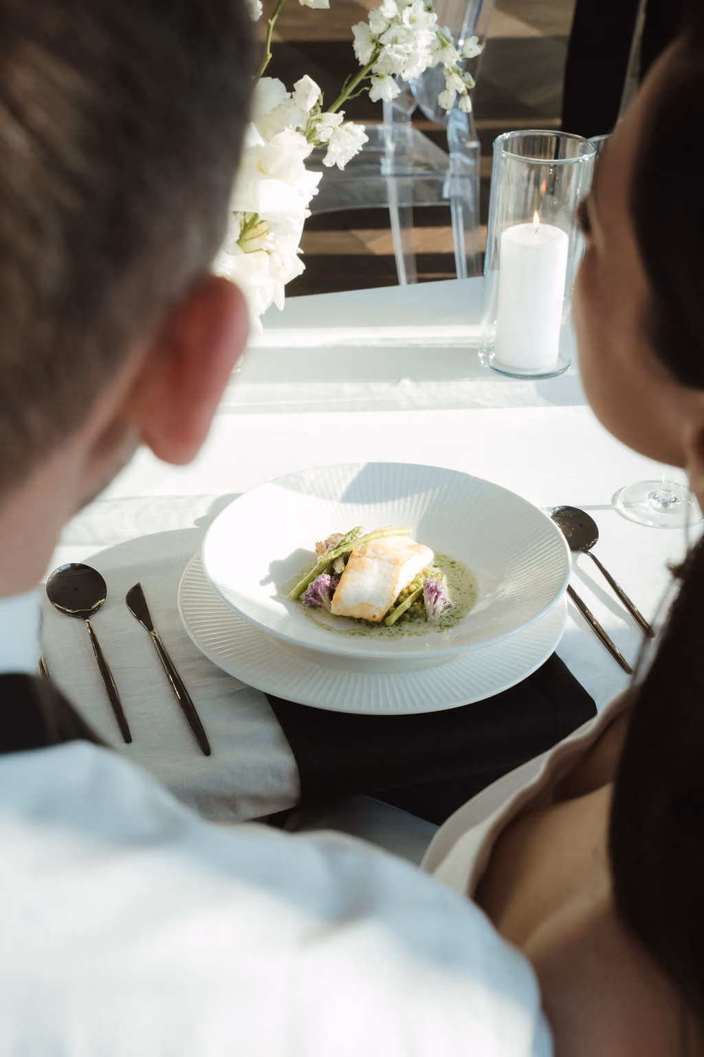 Ein Paar sitzt an einem gedeckten Tisch mit weißer Tischdecke und schwarzem Stoffserviette, vor ihnen ein Teller mit einem feinen Gericht aus Fischfilet, Spargel und grüner Sauce, daneben ein brennendes Glas-Kerzenlicht und weiße Blumen als Dekoration.