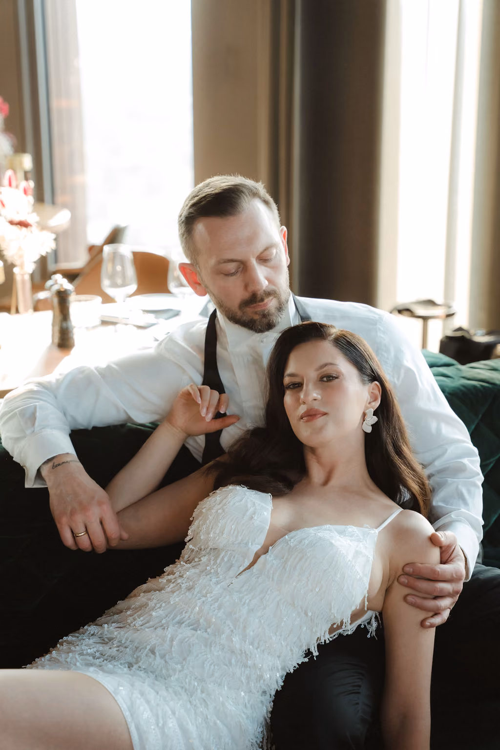 Ein Mann in weißem Hemd sitzt auf einem grünen Sofa und hält eine Frau in einem weißen, verzierten Hochzeitskleid, die sich entspannt an ihn lehnt.