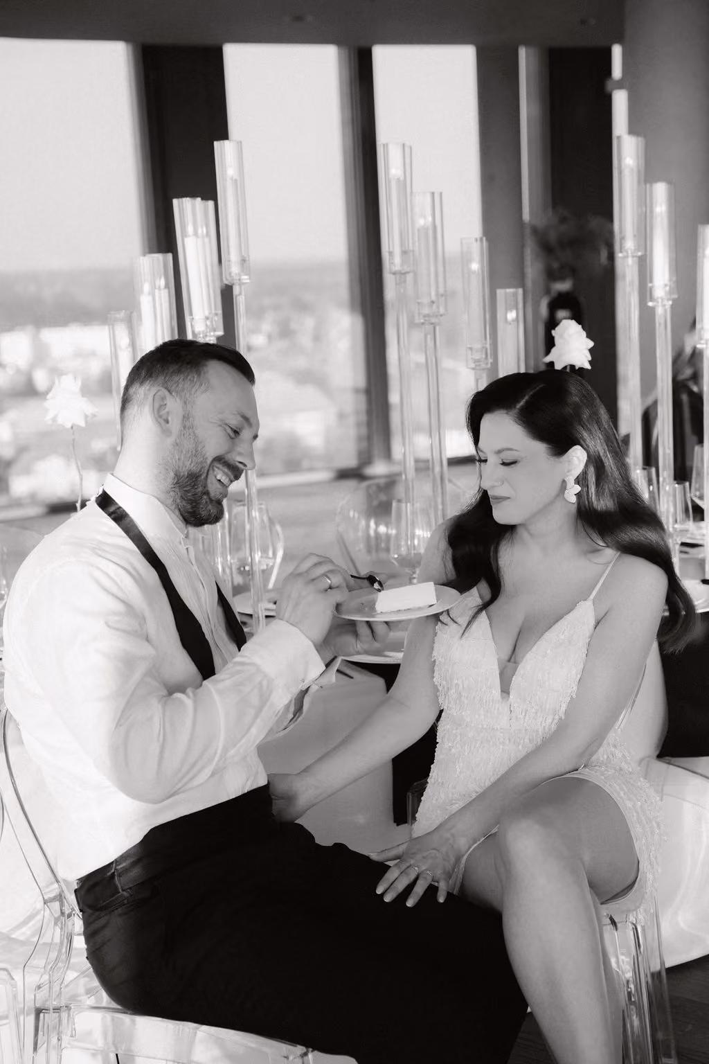 Schwarzweißfoto eines Paares bei einer Feier: Ein Mann in weißem Hemd und schwarzer Hose hält einen Teller mit einem Stück Kuchen und füttert lachend eine Frau in weißem Kleid, die ihm zugewandt sitzt.